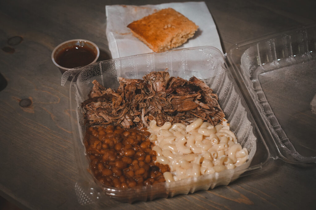 Clear plastic to-go container with pulled pork, Mac and cheese, and baked beans with a side of barbecue sauce and cornbread