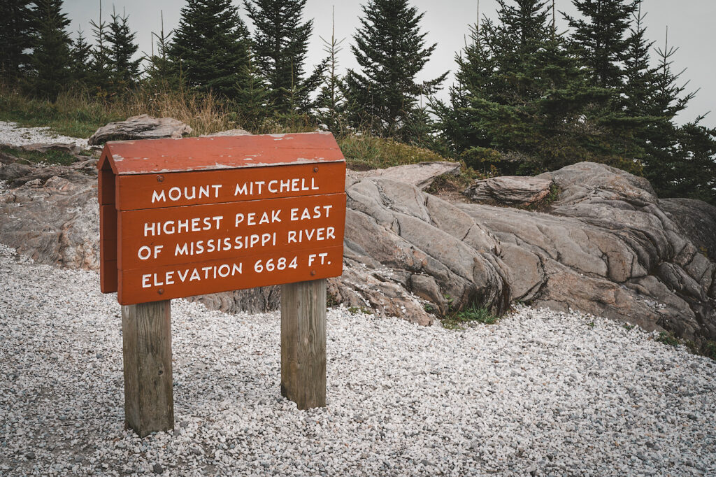 Red sign with white lettering that reads “Mount Mitchell Highest Peak East of Mississippi River” on it