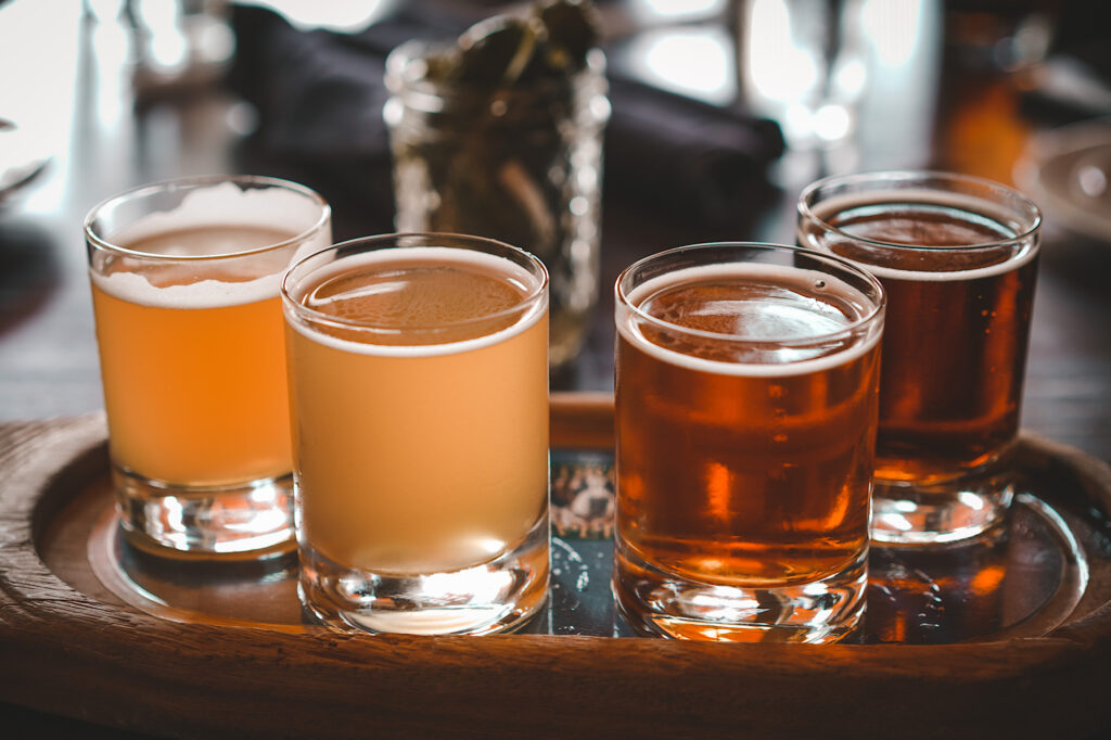 Flight of 4 beers sitting on wooden tray with light orange to brown in color order