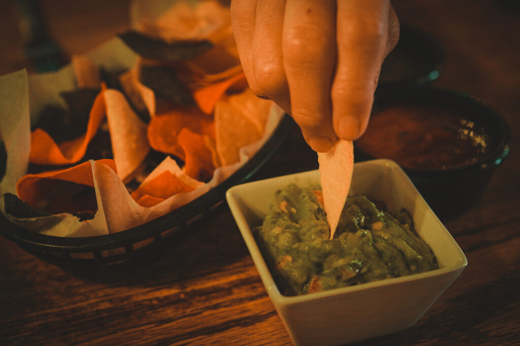 Hand dipping a chip in guacamole and multi-color tortilla chips on the side