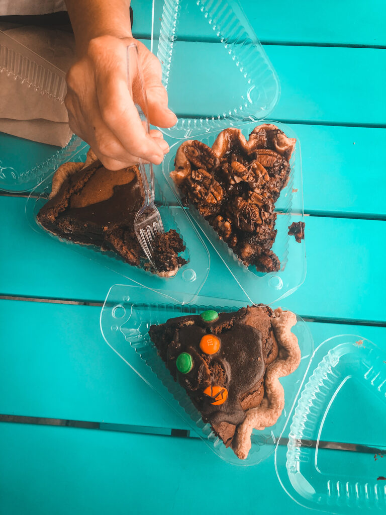 Bright blue table with one piece of chocolate pie with colored M&Ms, one piece of pecan pie, and one piece of chocolate pie with a hand getting a bite out with a fork